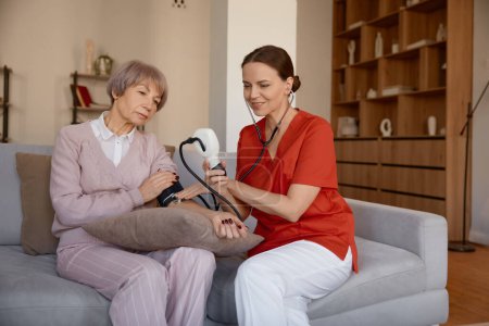 Foto de Trabajadora atenta de la salud usando tonómetro digital que toma la presión arterial de paciente mayor en el hogar de ancianos o apartamento de la casa de retiro. Servicio de chequeo cardiológico - Imagen libre de derechos