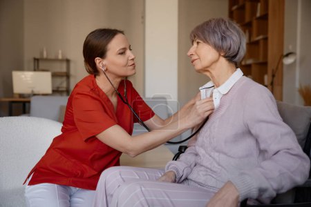 Téléchargez les photos : Femme médecin utilisant stéthoscope tout en examinant la femme âgée à la maison de retraite. Bilan médical professionnel pour les personnes âgées à la retraite - en image libre de droit