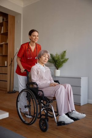 Photo for Woman caregiver preparing elderly lady in wheelchair for walk. Daily routine and everyday activities for old people in nursing home - Royalty Free Image