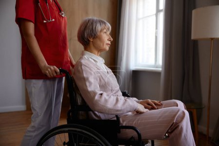 Téléchargez les photos : Femme âgée âgée en fauteuil roulant ayant une aide professionnelle à la maison de soins infirmiers ou de retraite. Vieillissement heureux avec support et concept de service de récupération - en image libre de droit