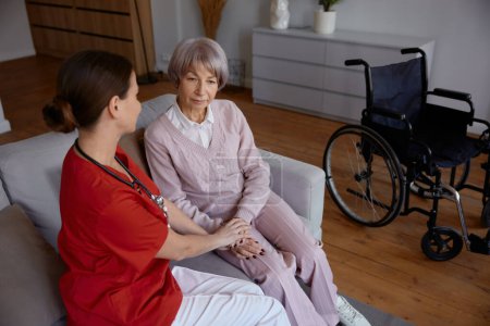 Téléchargez les photos : Médecin féminin parlant à sa patiente lors d'une visite à domicile. Femme médecin soutenant et aidant dame âgée en fauteuil roulant. Bonne retraite avec service professionnel - en image libre de droit