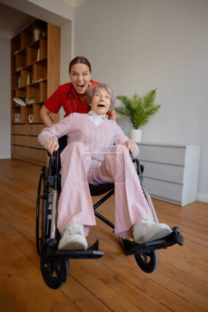 Téléchargez les photos : Femme folle soignante chevauchant une patiente âgée rapide en fauteuil roulant. Gardienne ludique appréciant le temps avec la vieille dame à la maison de soins infirmiers photo humoristique - en image libre de droit