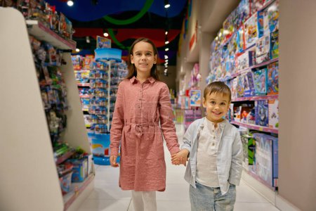 Téléchargez les photos : Petits enfants enchantés marchent parmi les vitrines avec des jouets en magasin. Adorable fille et garçon sélectionnant des jouets au supermarché ensemble - en image libre de droit