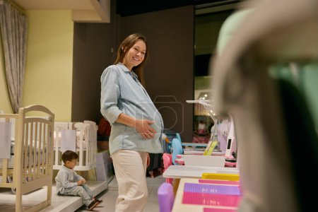 Téléchargez les photos : Mère enceinte souriante heureuse faisant du shopping en choisissant des meubles de sécurité pour le nouveau-né tandis que son petit fils assis se sent triste et ennuyeux - en image libre de droit
