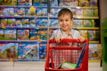 Téléchargez les photos : Portrait d'un petit garçon souriant se sentant heureux et excité avec chariot plein de jouets achetés debout sur l'affichage des jouets de jeu du supermarché. Vente rabais concept de publicité - en image libre de droit