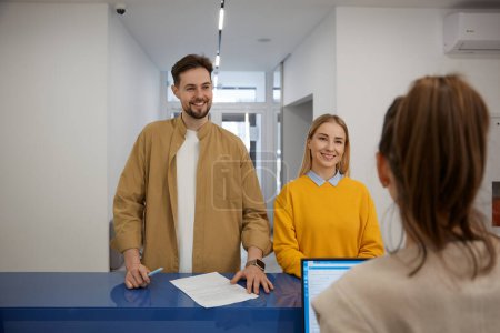 Happy man and woman couple signing paper documents at modern clinic reception desk. Young family filling questionnaire before doctor appointment while visiting hospital