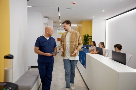 Photo for Man doctor discussing with young male patient results of routine checkup, diagnosis results using digital mobile tablet in corridor - Royalty Free Image