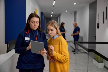 Photo for Doctor in uniform and patient talking discussing something looking at digital mobile tablet standing medical hospital corridor. Professional health discussion - Royalty Free Image