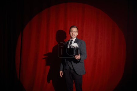 Photo for Elegant mysterious illusionist showing tricks with rabbit. Portrait of magician over red stage drapery. Wizard magic and fantasy illusion concept - Royalty Free Image