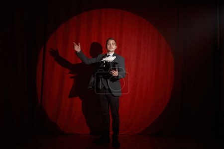 Foto de Elegante artista mago con pájaro paloma de pie sobre un paño rojo en la iluminación del centro de atención. Trucos de magia y espectáculo de ilusión milagrosa - Imagen libre de derechos