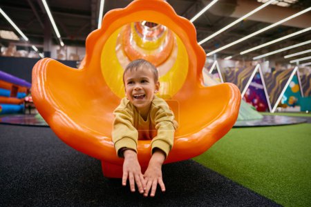 Cute little boy child having fun riding on slide at indoor playground. Portrait of pretty male kid feeling excitement and happiness at entertainment center
