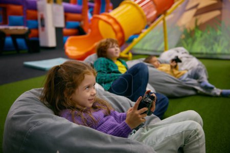 Téléchargez les photos : Petits enfants jouant à un jeu vidéo reposant sur des chaises douces après des jeux actifs dans le centre de divertissement - en image libre de droit