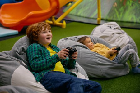 Téléchargez les photos : Portrait d'enfant garçon souriant heureux avec des joues roses jouant au jeu vidéo dans le centre des enfants. Temps de jeu - en image libre de droit