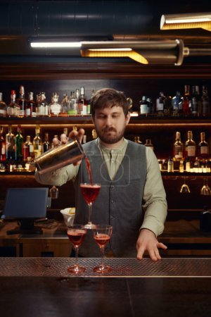 Téléchargez les photos : Amical barman souriant versant cocktail fraîchement préparé dans plusieurs verres sur le comptoir de bar en bois. Service professionnel pour groupe d'amis à la discothèque - en image libre de droit