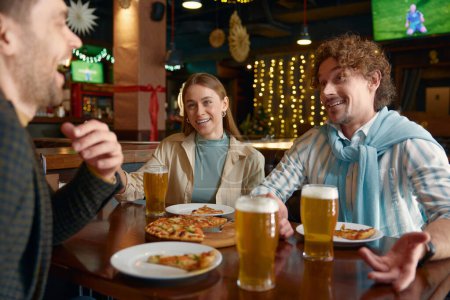 Téléchargez les photos : Les jeunes mangent une pizza en buvant de la bière pendant qu'ils se reposent au pub. Homme et femme couple rencontrer vieil ami mâle assis à la table avoir une conversation amusante, partager des moments intéressants de la vie - en image libre de droit