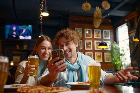 Foto de Jóvenes relajados mirando en el teléfono inteligente mientras descansan en el bar. Dos amigos felices se reunieron en un pub comiendo pizza, bebiendo cerveza, charlando y viendo videos divertidos en las redes sociales - Imagen libre de derechos