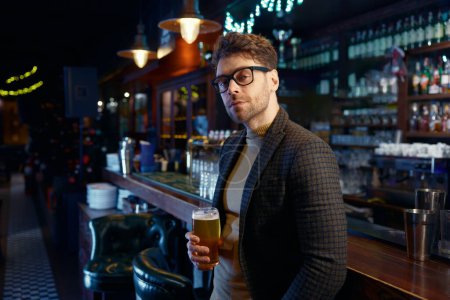 Téléchargez les photos : Portrait d'un homme d'affaires souriant et détendu tenant un verre de bière mousseuse froide dans un bar. Repos après le travail pendant, concept vendredi soir - en image libre de droit