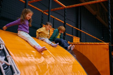 Téléchargez les photos : Joyeux petits enfants glissant de la surface en caoutchouc dans la piscine sèche en jouant sur le trampoline avec cube doux. aire de jeux intérieure loisirs amusants pour les enfants - en image libre de droit