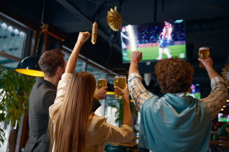 Glücklich aufgeregte Gruppe von Freunden schaut Fußballspiel im Fernsehen in der Kneipe. Blick zurück auf diverse junge Leute, die mit Bier über dem Kopf jubelnde Lieblingsfußballmannschaft beim Torjubel in die Höhe halten