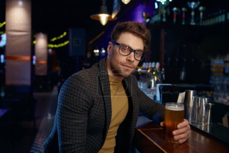 Téléchargez les photos : Pensif détendu souriant homme d'affaires tenant un verre de bière mousseuse froide dans le bar. Repos après le travail pendant, concept vendredi soir - en image libre de droit