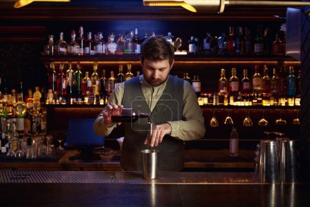 Téléchargez les photos : Barman concentré en uniforme versant la boisson alcoolisée des bouteilles dans le shaker en acier derrière le bar - en image libre de droit