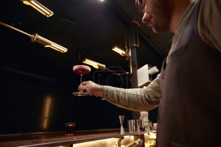 Téléchargez les photos : Satisfait barman se sentant excité de préparer le meilleur cocktail de bar. Barman tenant verre avec boisson décoré de pétales de fleurs debout au comptoir bar vue de côté - en image libre de droit