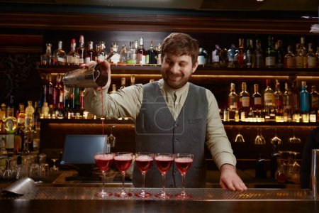 Téléchargez les photos : Amical barman souriant versant cocktail fraîchement préparé dans plusieurs verres sur le comptoir de bar en bois. Service professionnel pour groupe d'amis à la discothèque - en image libre de droit