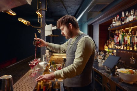 Téléchargez les photos : Barman souriant versant un cocktail fraîchement préparé dans plusieurs verres sur le comptoir du bar en bois. Service professionnel pour groupe d'amis à la discothèque - en image libre de droit