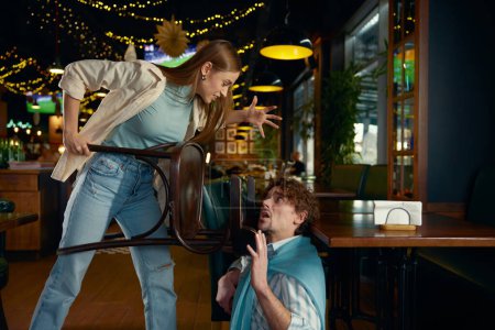 Photo for Emotional fight and conflict between drunk man and woman couple in bar. Furious young girlfriend attacking fighting boyfriend pushing him with chair - Royalty Free Image