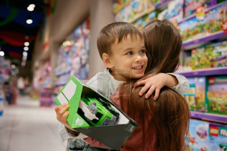 Porträt eines glücklichen Sohnes, der seine Mutter im Spielzeugladen umarmt. Kleines Kind, das dankbar für gekauftes Spielzeug ist. Familieneinkaufskonzept