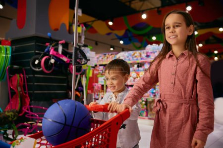 Alegre hermanita y hermano haciendo compras en tienda de juguetes. Retrato de niña feliz y niños hermanos empujando carro con un montón de juguetes