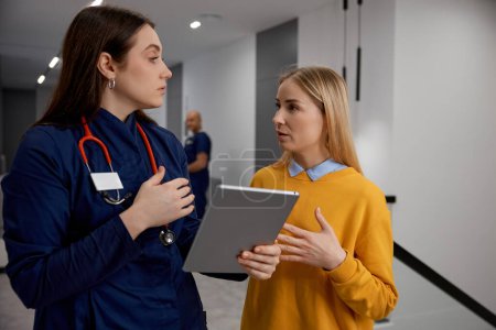 Photo for Two young woman, doctor in uniform and patient talking discussing something looking at digital mobile tablet standing medical hospital corridor. Professional health discussion - Royalty Free Image