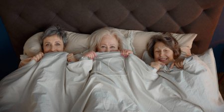 Photo for Cute funny elderly woman friends lying in bed under blanket enjoying free time celebrating party together. Happiness, friendship and retirement - Royalty Free Image