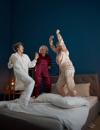 Foto de Alegres amigas ancianas positivas usando ropa de dormir escuchando música bailando en la cama. Amistad, unión y despedida de soltera en la pensión - Imagen libre de derechos