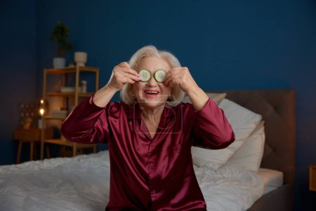 Photo for Happy smiling aged elderly woman in pajamas applying cucumber mask on eyes. Senior beauty and skin care procedures at home - Royalty Free Image