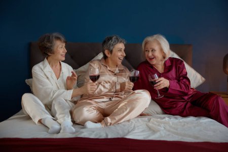 Foto de Hermosas amigas mayores en pijama bebiendo vino disfrutando de un divertido tiempo de retiro juntas. Saludable atractivas mujeres maduras que hablan chismes reunión en la fiesta de pijamas en casa - Imagen libre de derechos