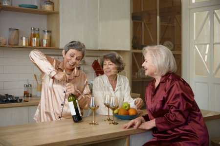 Téléchargez les photos : Dames à la retraite excitées en pyjama déroulant bouteille de vin à la maison cuisine. Trois sœurs âgées célébrant des vacances ensemble boire de l'alcool se sentent bien dans une atmosphère idyllique et confortable - en image libre de droit