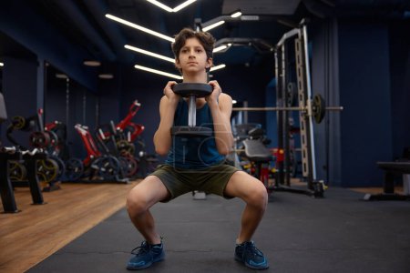 Foto de Guapo chico joven deportivo disfrutando de la actividad de fitness en el gimnasio. Niño preadolescente haciendo ejercicio en cuclillas con pesadas mancuernas bombeando sus músculos - Imagen libre de derechos