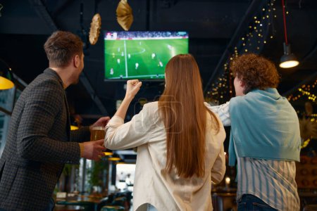 Glücklich aufgeregte Gruppe von Freunden schaut Fußballspiel im Fernsehen in der Kneipe. Blick zurück auf diverse junge Leute, die mit Bier über dem Kopf jubelnde Lieblingsfußballmannschaft beim Torjubel in die Höhe halten