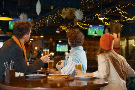 Group of friends watching football match on tv in pub. Back view on diverse young people holding glasses with beer raised over head cheering favorite soccer team celebrating goal in gate