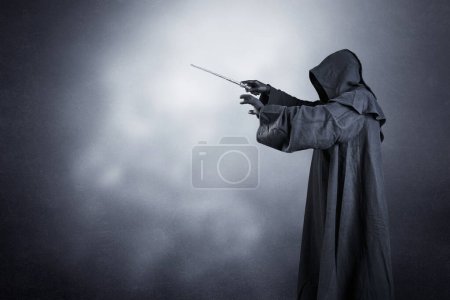 Téléchargez les photos : Magicien avec cape à capuchon et baguette magique sur fond sombre brumeux - en image libre de droit
