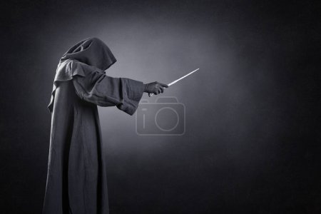 Foto de Asistente con capa encapuchada y varita mágica sobre fondo oscuro brumoso - Imagen libre de derechos
