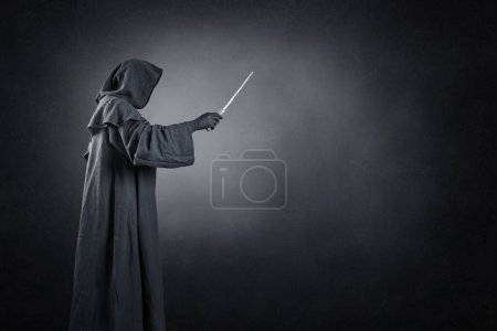 Foto de Asistente con capa encapuchada y varita mágica sobre fondo oscuro brumoso - Imagen libre de derechos
