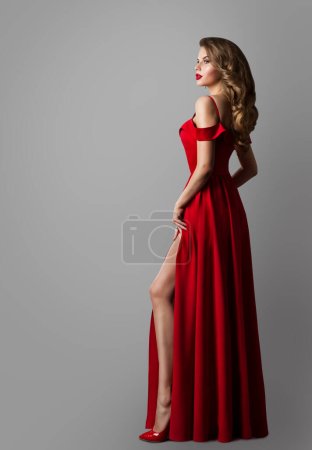Foto de Modelo de moda en vestido largo rojo. Mujer elegante en vestido de noche sobre gris. Sexy Girl mostrando Leg in Slit. Mujer Wavy Curly Peinado. Lady Style Ropa de vacaciones - Imagen libre de derechos