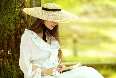 Foto de Chica romántica en vestido blanco y sombrero de sol de paja lectura libro sentado bajo el árbol. Mujer joven estudiando en Spring Park. Old Fashioned elegante dama fuera - Imagen libre de derechos