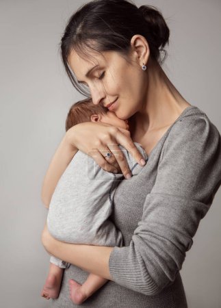 Happy Mother holding Baby in Arms. Newborn Health Care and Development. Beautiful Mom embracing little Son over Gray Background. Smiling Woman enjoying parenting