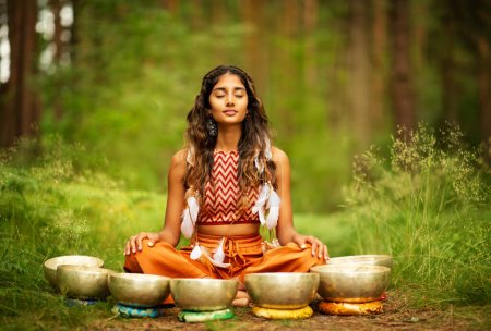 Photo for Indian Woman meditating with Tibetan Singing Bowls Outdoors. Yoga Practice in Forest. Spiritual Sound Healing Therapy. Peaceful Nature Relaxation - Royalty Free Image