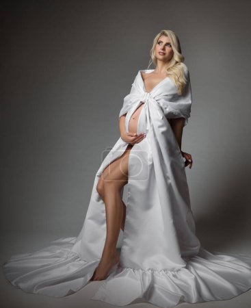 Foto de Mujer embarazada en vestido de seda blanca abrazando vientre. Hermosa madre en vestido largo soñando con fondo gris oscuro. Retrato artístico del embarazo - Imagen libre de derechos