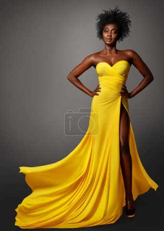 Foto de Moda mujer de piel oscura en vestido de seda amarillo volando sobre gris. Modelo Africano con peinado afro rizado en vestido de noche largo. Elegante retrato de dama - Imagen libre de derechos