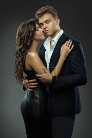 Photo for Handsome Man in Suit embracing Beautiful Girl side view over Gray. Fashion Couple. Couple in Love dating. Elegant Gentlemen and Lady in Black cocktail Dress - Royalty Free Image